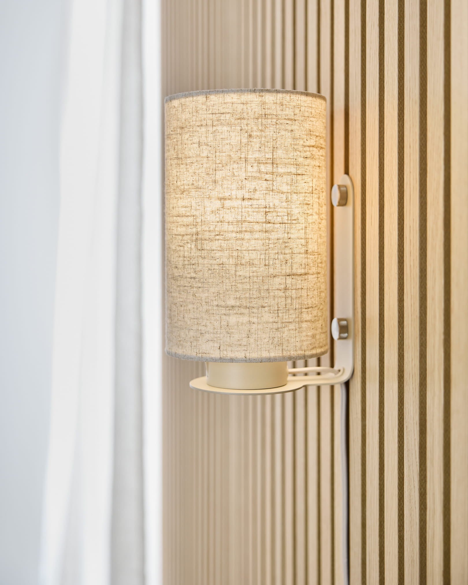 The Feya Wall Lamp in Beige by WoodUpp casts warm light onto a beige Akupanel acoustic slat wall, positioned next to a white curtain and linen-cotton shade.